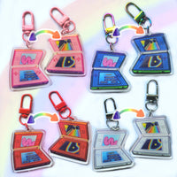Eight retro handheld game console keychains arranged in a grid on a white background with a soft pastel rainbow glow. Each keychain features a translucent acrylic game console charm with a colorful screen graphic and a metal clasp in different color combinations: pink, blue, red, and silver.