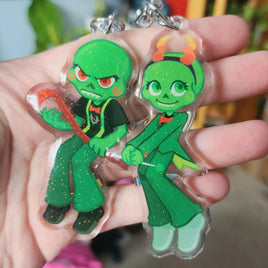 Two green cartoon character keychains being held in a hand against a blurred background.