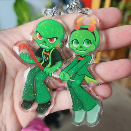 Two green cartoon character keychains being held in a hand against a blurred background.
