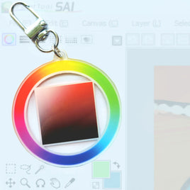 A circular acrylic keychain with a rainbow gradient ring and a central square color swatch, attached to a silver metal clasp. The background is a blurred digital color picker interface.