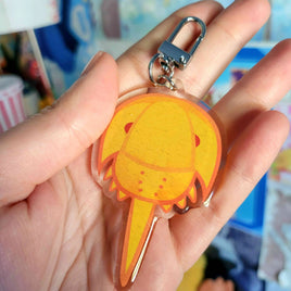 A hand holding a clear acrylic keychain charm of a horseshoe crab with a silver metal clasp, against a blurred indoor background.