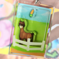 A clear acrylic keychain or charm featuring an anime-style brown horse character standing in a green field with a white fence, held in a hand against a soft pastel blurred background.
