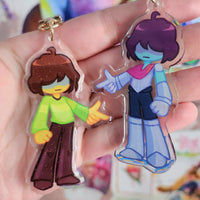 Two anime-style acrylic keychains held in a hand. One keychain features a character with brown hair and a green shirt, and the other features a character with purple hair, blue skin, and a pink and blue outfit.