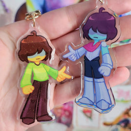 Two anime-style acrylic keychains held in a hand. One keychain features a character with brown hair and a green shirt, and the other features a character with purple hair, blue skin, and a pink and blue outfit.