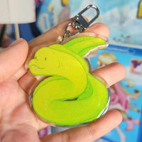 A hand holding a translucent acrylic keychain charm of a moray eel, with a silver metal clasp attached. The background is a blurred blue bedroom.