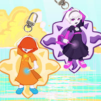 Two anime-style keychain charms with metal swivel clips, shown on a bright pastel background. One charm is a hooded figure in orange with blue shoes and a magic wand omitting a blue glow